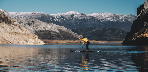 a woman with yellow plumber paddle surfing on a lake in a snowy mountain landscape
