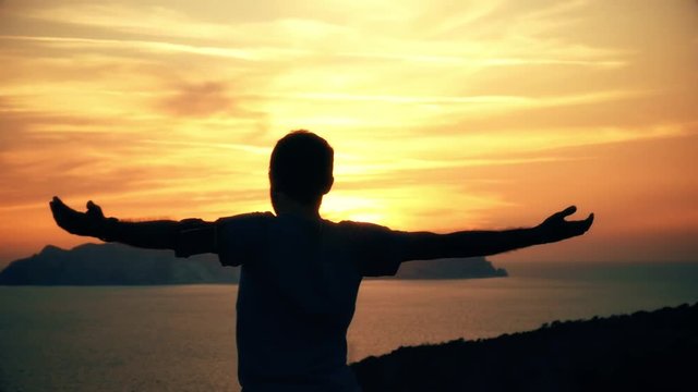Silhouette of active young man stretching doing yoga exercise on top of a mountain cliff on ponza island at sunset with the sea landscape on the background slow motion