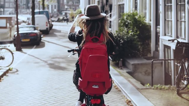 Tourist girl riding bicycle in sunny city street. Turning right. European girl in hat on a bike. Back view slow motion.