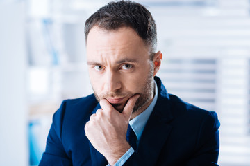 Gloomy mood. Young experienced elegant businessman spending time in a big comfortable office with his hand touching his face and looking upset