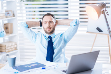 Unpredictable situation. Worried experienced skilled manager sitting in an armchair with the arms behind his head and looking upset while thinking about an unpredictable unpleasant situation at work