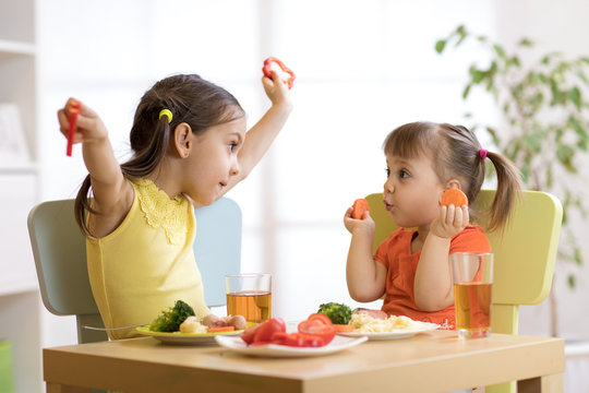 Cute smiling child and toddler girls playing and eating spaghetti with vegetables for healthy lunch sitting in a white sunny kitchen with big window