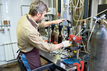 Bearded middle-aged worker wearing apron and gloves operating beer bottling equipment while standing at spacious modern brewery