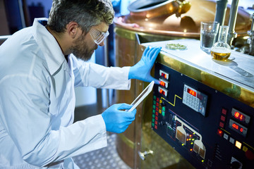 Middle-aged brewer wearing white coat and protective goggles adjusting machine with help of operation manual presented on screen of digital tablet, interior of modern brewery on background