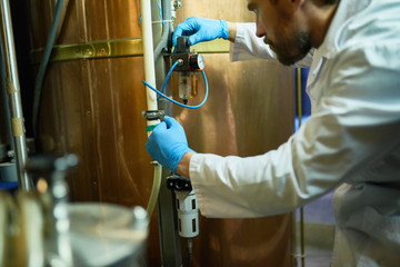 Confident brewer wearing white coat and rubber gloves adjusting pressure in cylindrical beer maturation tank while working at modern brewing plant