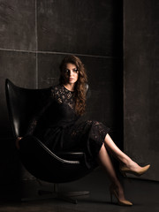 Beautiful young woman in a black lace dress sitting in a chair. Dark portrait