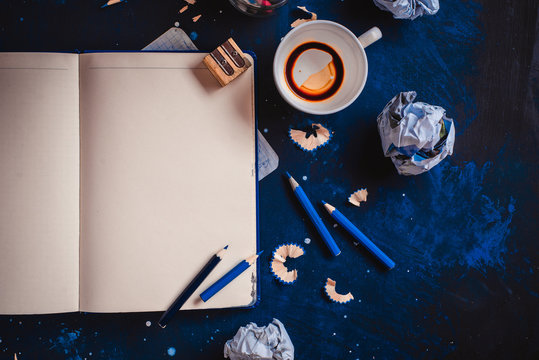Open notepad with blank pages, crumpled paper balls, pencils, notepads and empty coffee cups on a dark background. Still life with writer workplace. Creative writing concept.