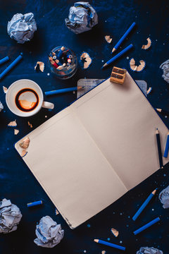 Open notepad with blank pages, crumpled paper balls, pencils, notepads and empty coffee cups on a dark background. Still life with writer workplace. Creative writing concept.