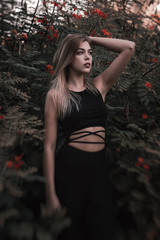 Vertical photo of a female blond model in black trendy jumpsuit standin near green bush with red flowers