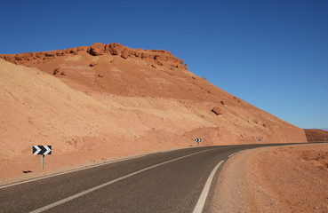 Widok asfaltowej drogi biegnącej przez skalisty teren pustynny, droga skręca w prawo, po lewej skaliste wzgórze w kolorze piasku, błękitne czyste niebo - obrazy, fototapety, plakaty