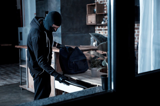 Professional burglar. Violent professional masked burglar wearing a black uniform and gloves and stealing a laptop from the table and holding a bag
