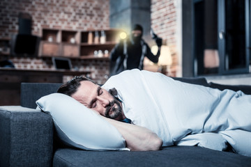 Sleeping. Peaceful dark-haired bearded man lying in bed and sleeping and the criminal with a torch...