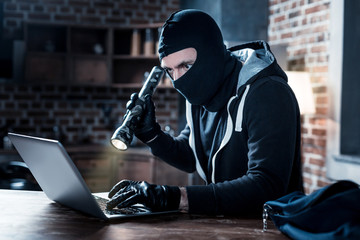 Computer hacker. Serious professional masked computer hacker sitting at the table and holding a...