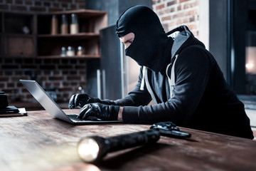Torch and gun. Concentrated trained masked computer hacker sitting at the table and stealing data...