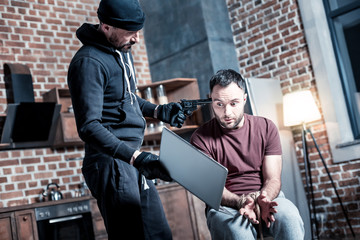 Transfering money. Panic intimidated bearded man sitting with his hands tied while a killer standing near him and holding a gun and a laptop and demanding transferring money