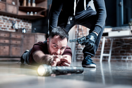 Reaching for torch. Scared frightened bearded man lying on the floor and having his hands tied and reaching for a torch while a killer standing behind him and holding a gun at his head