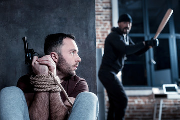 Hiding himself. Intimidated shocked bearded young man holding a gun and sitting with his hands tied...