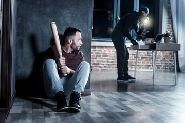 Hiding. Frightened dark-haired bearded man hiding and holding a stick while a robber holding a...