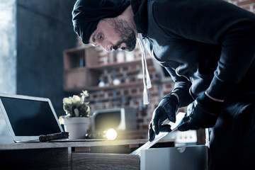 Documents. Determined experienced bearded burglar standing near the table and looking for documents in it and having a knife and torch with him