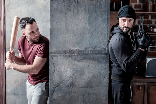 Fighting. Determined Dark-haired Beaded Man Holding A Stick And Hiding From The Killer Standing Behind The Wall And Holding A Gun