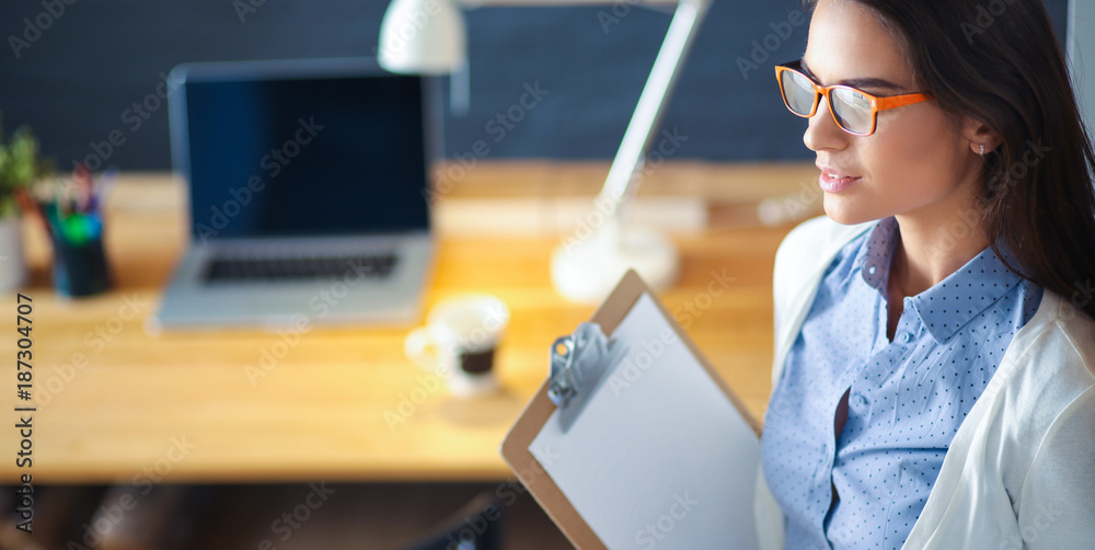 Wall mural young woman standing near desk with laptop holding folder and cup of coffee