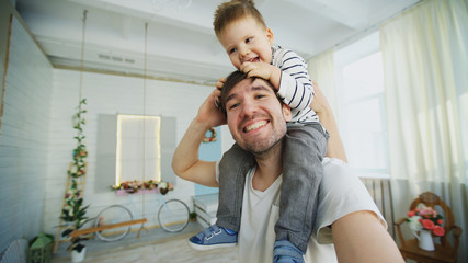 Happy father carrying his smiling son on neck and making selfie on smartphone in bedroom