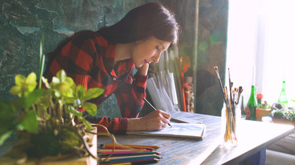 Young woman artist painting sketch on paper notebook with pencil. Bright sun flare from window