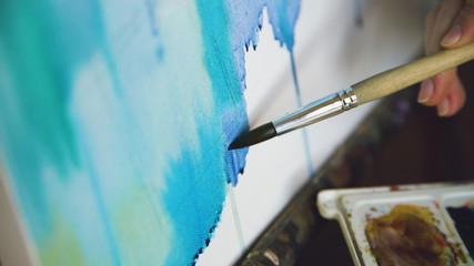 Naklejka premium Young woman artist draw pictrure with watercolor paints and brush on easel canvas closeup hand