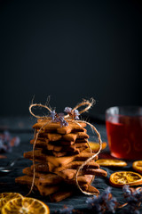 Tied stack of star shaped gingerbread cookies and a glass cup of red tea. Decorated with dried violet flowers, and dried orange slices, dark background.