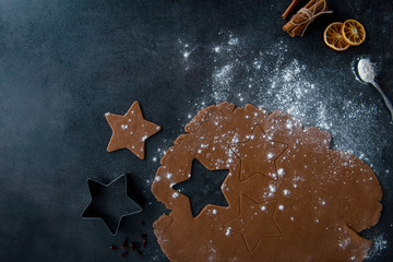 Christmas gingerbread star cookie cutter, gingerbread dough with cookies forms cutout, teaspoon of flour, cinnamon sticks, dried orange slices and rolling pin, dark background, top view.