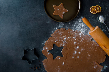 Christmas gingerbread star cookie cutter, gingerbread dough with cookie cutout, star shaped cookie on plate, teaspoon of flour, dried orange slices and rolling pin, dark background, top view.