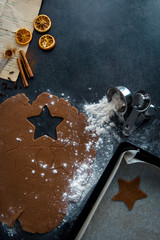 Christmas gingerbread star cookie cutout on gingerbread dough with flour, measuring cups with flour and cookie in sheet pan, cinnamon sticks and orange slices. Dark background, flat lay.