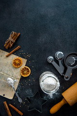 Food preparation. Old recipe, measuring cups, cookie cutter, rolling pin, mason jar with flour and ingredients (cinnamon sticks, dried orange slices) on dark modern table from above. Top view.