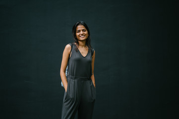 Portrait of cheerful and smiling young Indian lady with a backpack in a grey jumpsuit against a dark wall