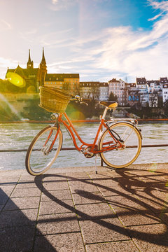 city bicycle with basket on the steering wheel of red color on the quay near the river Rhine in Switzerland against the backdrop of old city and authentic houses and cathedral on sunny day in winter