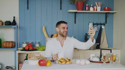 Cheerful young man using smartphone for online video chat with girlfriend while have breakfast in...
