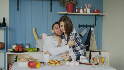 Attractive couple meet in the kitchen at breakfast time and using smartphone for social media surfing at home