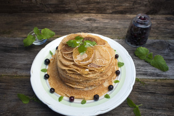 Pancakes are located in a plate on an old wooden table decorated with berries and mint leaves. Pancakes are watered with syrup.