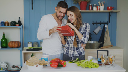 Happy cheerful man surprising his girlfriend with a gift at home in the kitchen while she cooking breakfast