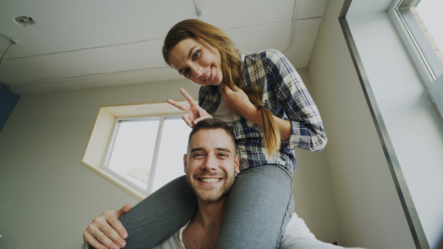 POV of happy smiling and beautiful couple take selfie portrait on smartphone camera while girl sitting on man's neck at home