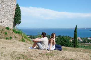 Handsome man and a young woman sitting next to him on the dried grass near an old castle on a hill overlooking the sea and the blue sky