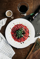 beef carpaccio with arugula on a white plate with a glass of red wine and pine nuts on a dark background