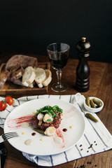 Beef tartare with quail eggs, cornichons and arugula, with bread and a glass of red wine on a dark wooden table