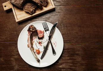 Dirty plate with eaten food, bone and folded cutlery with a bread board on a wooden table - 187329120