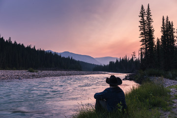 Cowboy sitting by a raging river at sunset. The gorgeous colors of an Alberta sunset are displayed as the green grass grows and the river rushes by. The man sits with a denim jacket and cowboy hat.