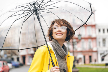 Amazing portrait of happy woman in yellow raincoat walking in city under transparent umbrella...