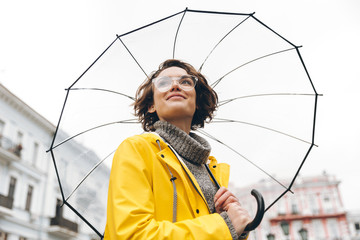 View from beneath of positive woman in yellow raincoat and glasses standing in street under big...