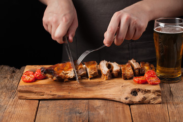 The chef cuts it with a sharp knife ready to eat pork ribs, lying on an old wooden table. A man prepares a snack to beer on a black background with copy space
