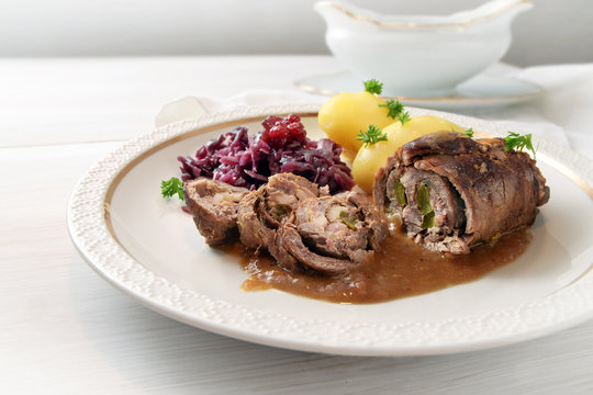 beef roulade with red cabbage and potatoes, german meat roll stuffed with cucumbers, bacon and onions on an elegant white gold rim plate on a light wooden table, copy space