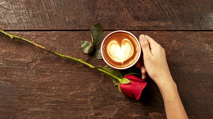 Hand of a young woman holding a red coffee cup on an old wooden desk with roses, conveying love.
